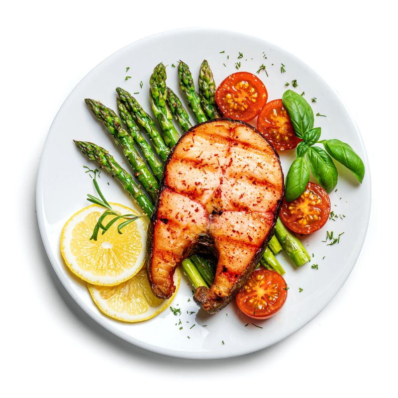 Grilled salmon steak with green asparagus, tomatoes and lemon slices served on white plate