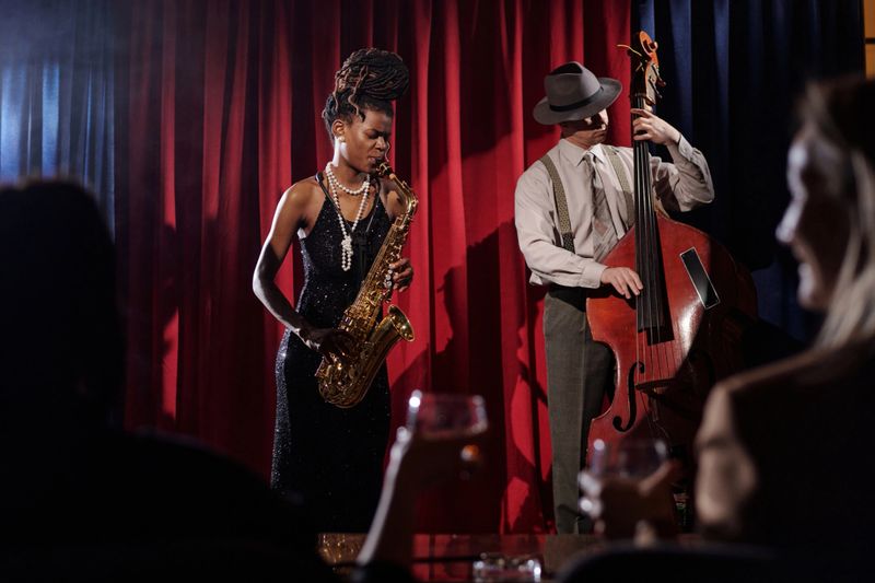 Black young adult woman playing saxophone and Caucasian middle aged man playing double bass performing jazz music on stage with red and blue curtain, audience watching in foreground