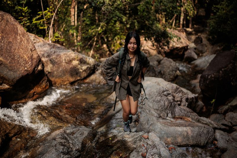 A young woman is climbing uphill, exploring the forest of a national park in Koh Phangan, Thailand, with a backpack on her back, enjoying the view offered by the vast forest she is exploring.