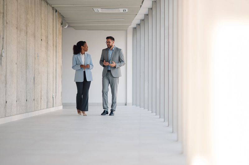 Businessman and businesswoman walking and engaging in conversation while navigating a modern office hallway, showcasing collaboration and teamwork