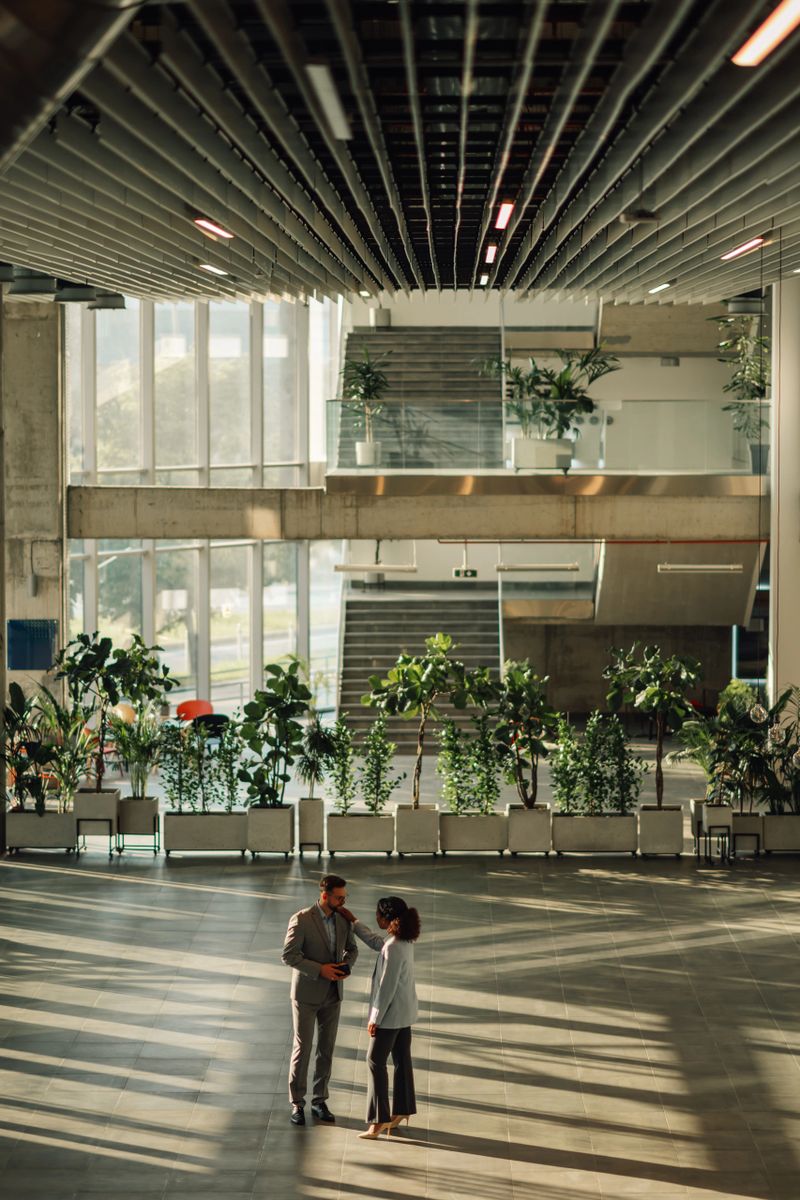 Two businesspeople engaging in conversation within a modern office lobby, surrounded by lush plants and sleek architectural design
