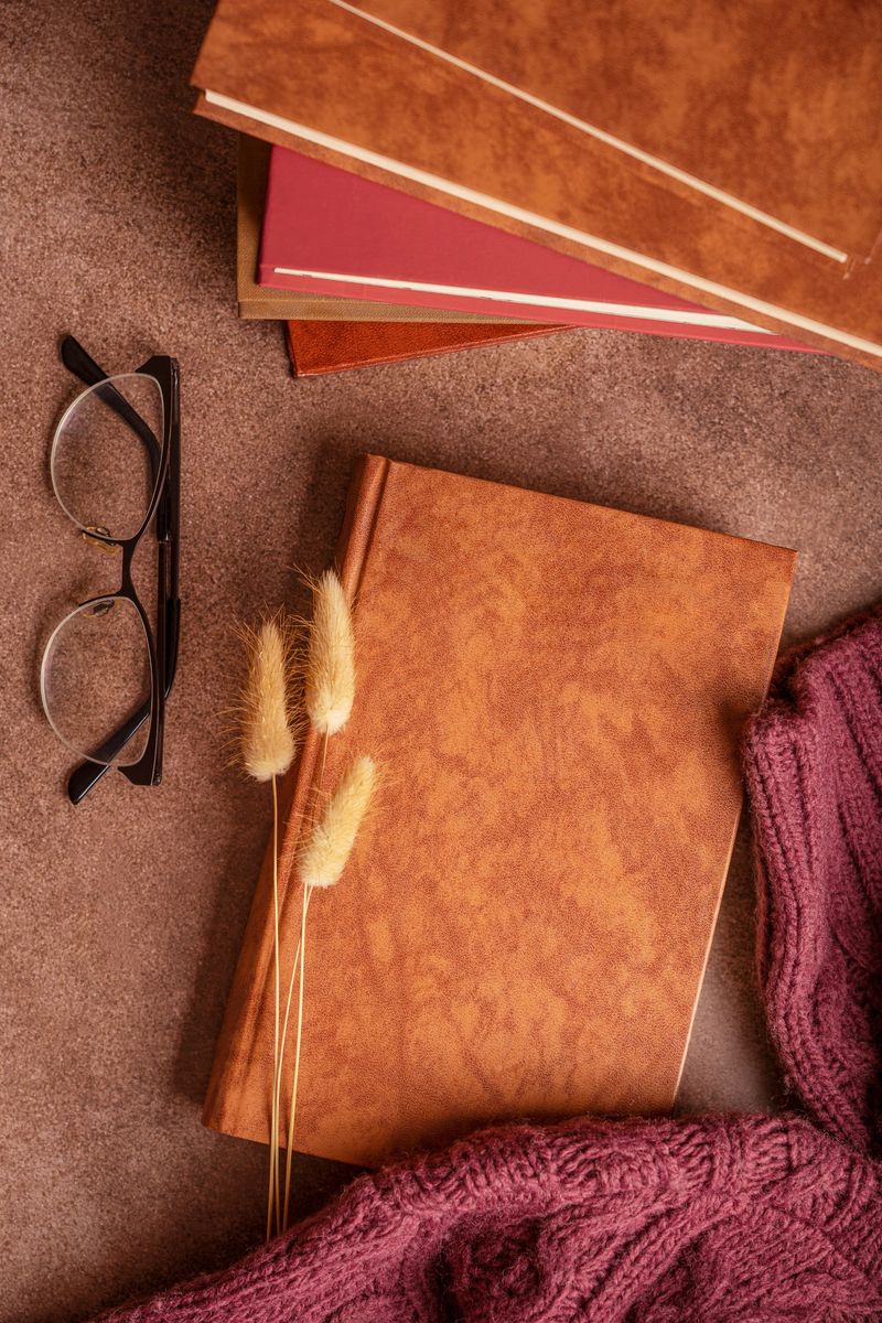 Top view flat lay of books, eyeglasses, and dried flowers on dark brown table with warm autumn textile at reading corner indoors at home or library prepared for cozy comfortable evening or education