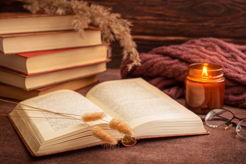 Open book with dried autumn plants on dark brown table with glowing candle light, glasses and warm cozy textile cloth against wooden wall at reading corner during comfortable home leisure evening