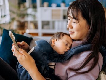 Woman holding a sleeping baby and looking at a smartphone.