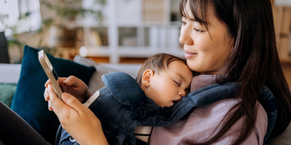 Mother holding sleeping baby while using phone.