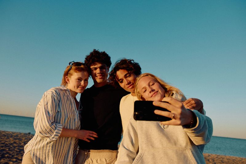 Four young friends, two females and two males, take a cheerful selfie on a sandy beach at sunset, with the lake and blue sky forming a beautiful backdrop.