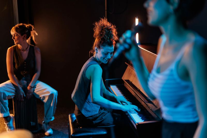 A vibrant all-female trio performs with joy and harmony — one sings with a microphone in hand, another plays the piano, while the third keeps the rhythm on the cajon. Each of them wears a beautiful smile, radiating connection, confidence, and the pure happiness of making music together.