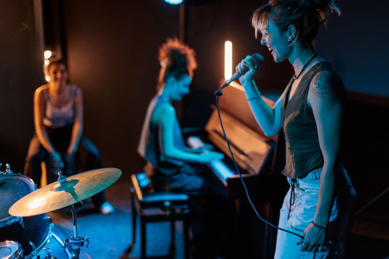 In the spotlight stands a beautiful woman singing passionately with a microphone in her hands, her expression confident and full of emotion. Behind her, two fellow band member, one on piano and the other on cajon.