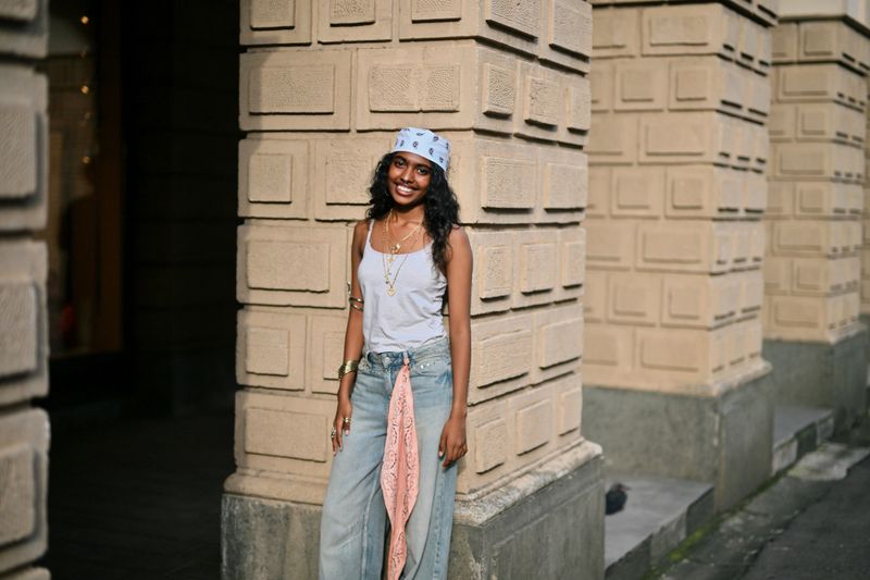 A cheerful teenage girl smiles brightly while standing outside an urban building with textured stone pillars. She wears a casual white tank top, loose blue jeans, layered gold jewelry, and a light blue bandana, giving a youthful vibe.