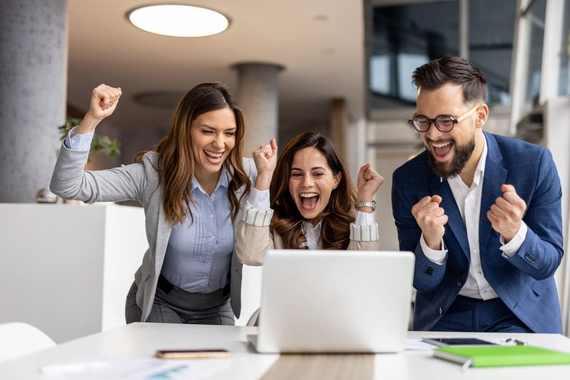 Three enthusiastic businesspeople are cheering and raising their fists in front of a laptop, expressing their excitement over a positive outcome in a modern office setting
