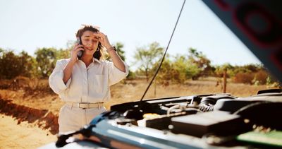 Worried woman calls for help next to a broken-down car with open hood.