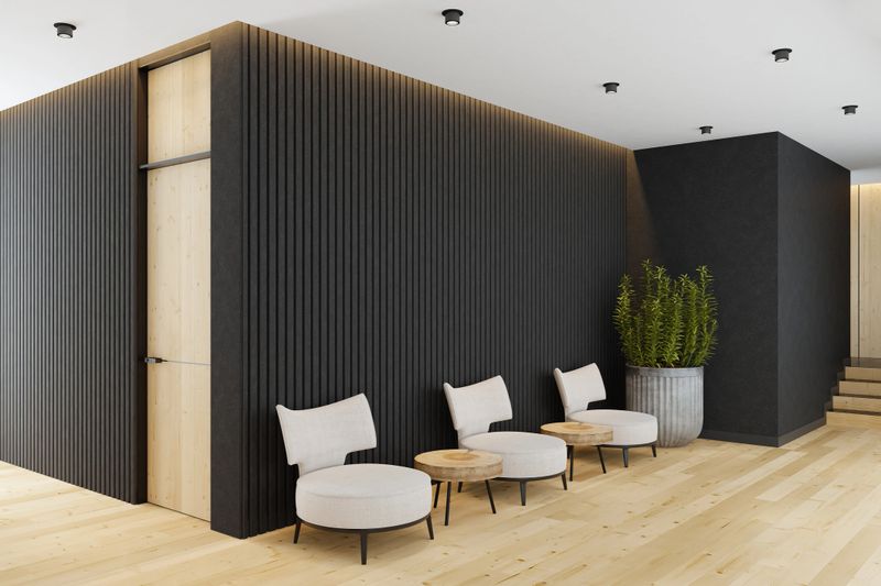 Spacious modern entrance hall featuring dark stone walls that create a striking, textured backdrop paired with long matte pine laminate parquet flooring in a natural finish. The combination of rugged stone and warm wood tones delivers a balanced Scandinavian aesthetic with a contemporary edge. Three armchairs with rustic wooden stump side tables add artistic detail and cozy seating. The white ceiling is fitted with sleek black tubular spotlights, providing clean, focused lighting. Perfect for offices, medical or dental clinics, reception areas, waiting rooms, and creative work spaces, this versatile interior blends natural materials and modern design for a professional yet inviting atmosphere.