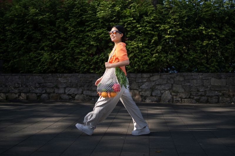 Young woman walking outdoors, carrying a reusable mesh bag filled with fresh vegetables and fruit
