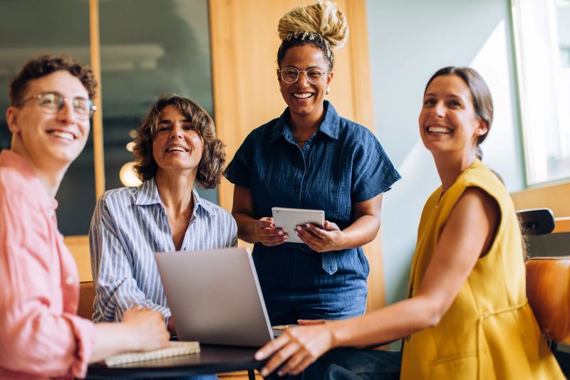 Group of coworkers gathered around a table, smiling and working on laptops and tablets, showcasing teamwork and friendly interaction in a modern office setting.