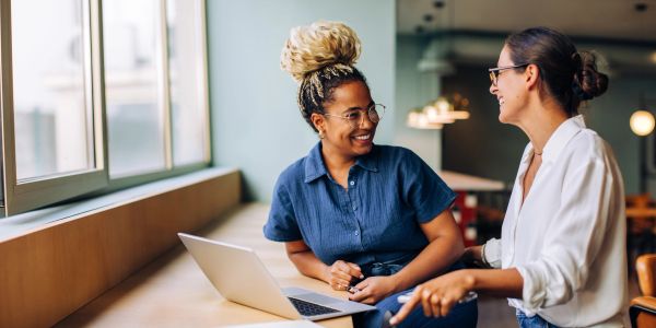 Two women happily discussing work at a laptop in a modern office.