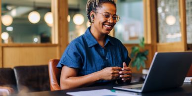 Smiling woman video chatting on a laptop in a cozy office.