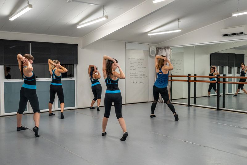 A dance teacher leads four students through a choreographed routine in a mirrored studio