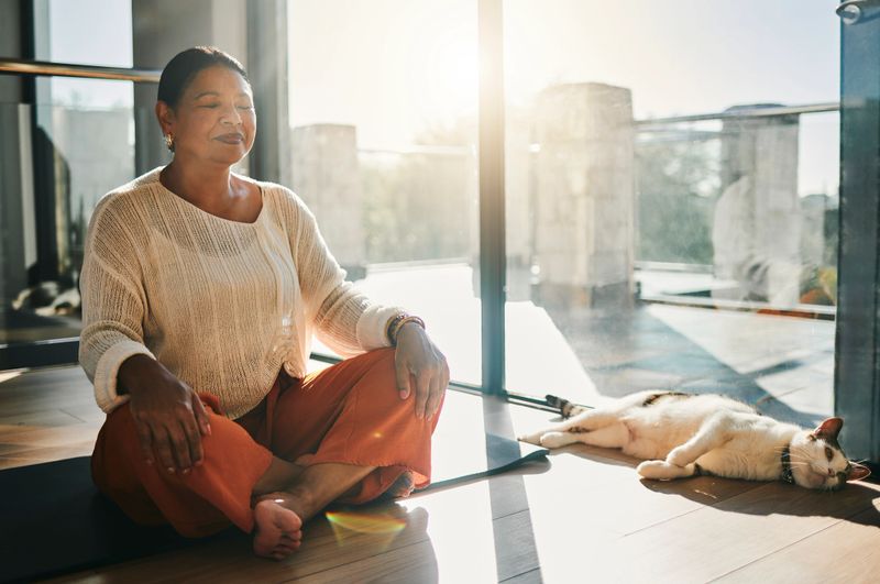 A serene woman of diverse ethnicity meditates in a warm, sunlit room, accompanied by a relaxed cat lying peacefully on the floor.