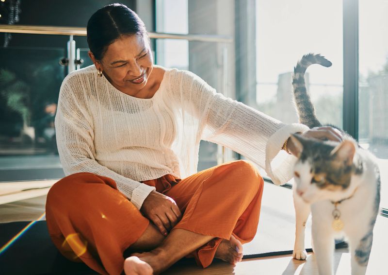 A cheerful woman wearing orange pants and a white sweater sits on the floor, petting a cat in a brightly lit, modern setting.