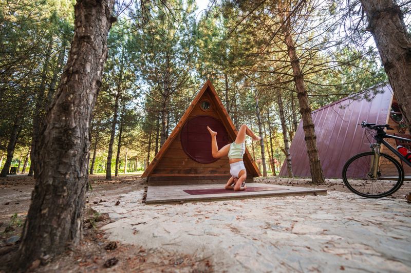A woman practicing exercises on her yoga mat in front of a triangular wooden public cabin in the forest, enjoying a peaceful yoga session amidst natural serenity.