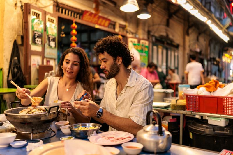 Sitting close, a couple dips ingredients into a steaming hot pot, enjoying the warmth and flavors of the meal together. Chinatown in Bangkok, Thailand.