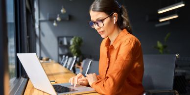 Woman working on laptop with glasses and earbuds.