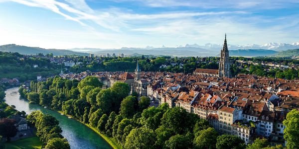 A picturesque European town with a river, lush greenery, and historic buildings under a clear sky.