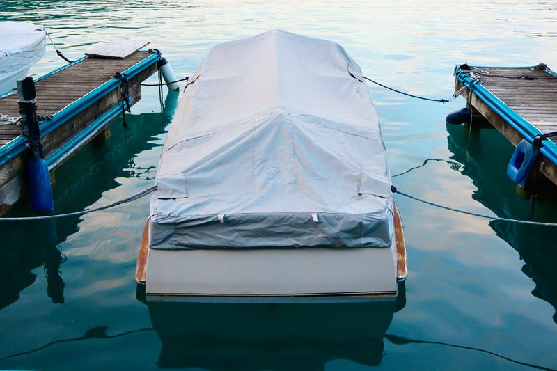 Small motorboat covered with a protective gray tarp, docked between two wooden piers on calm turquoise water. Peaceful marina scene with reflections.