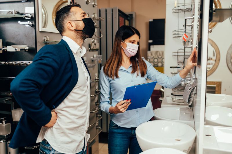 Shop assistant showing bathroom fixtures to customer wearing protective face masks in a modern showroom