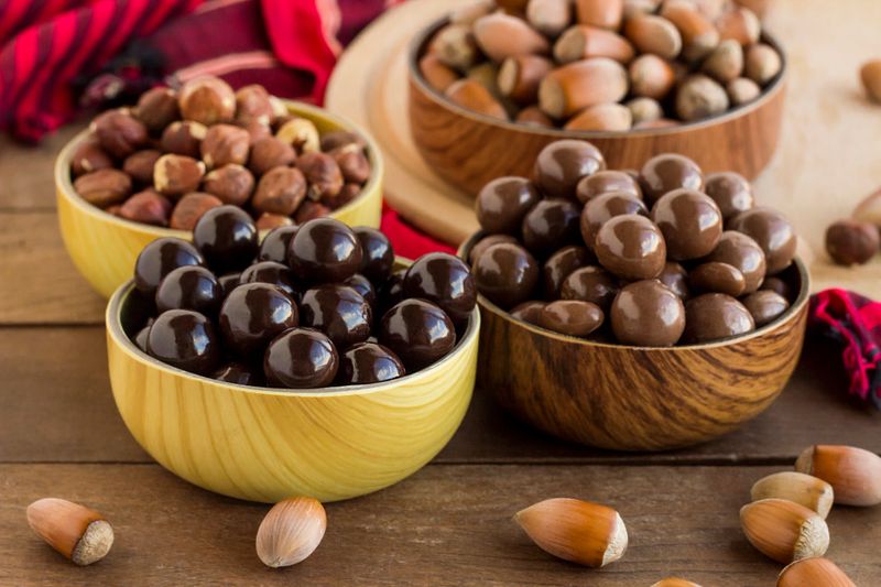 Milky and dark chocolate covered hazelnuts in bamboo bowls on wooden table with natural hazelnuts.