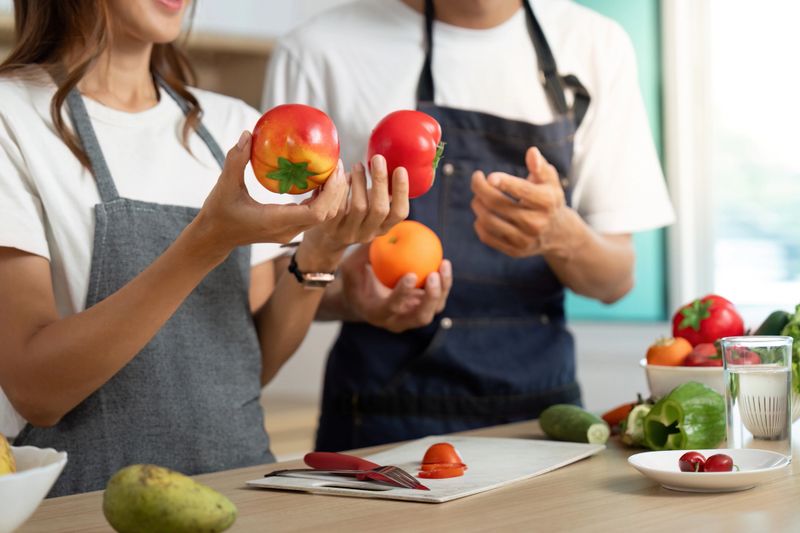 A couple shares a fun cooking moment, holding fresh tomatoes and discussing their meal prep in a bright kitchen.