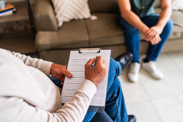 Person writing on clipboard with another person sitting in background.