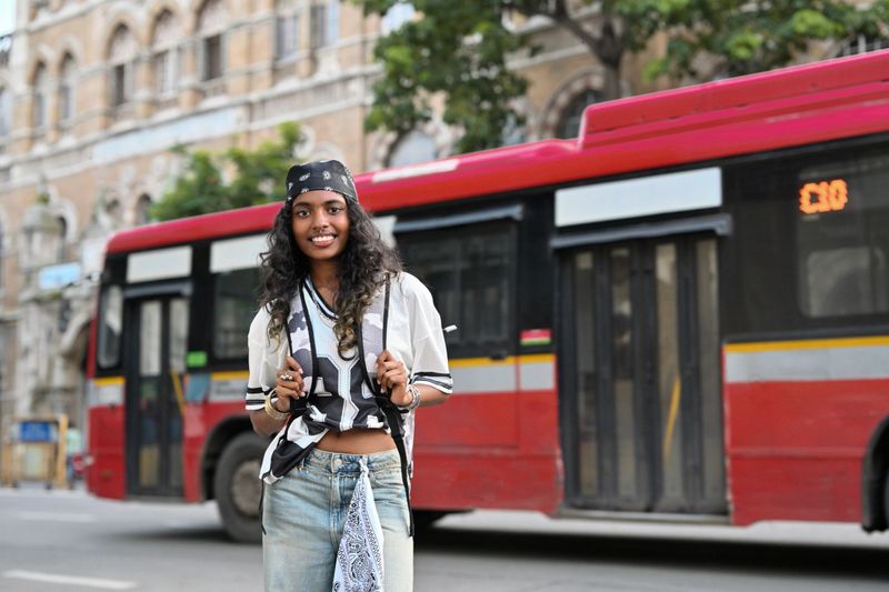 A smiling teenage girl with a confident look smiling at camera in front of a red city bus on an urban street. Her casual and sporty outfit reflects a cool, youthful vibe against a bustling city backdrop.