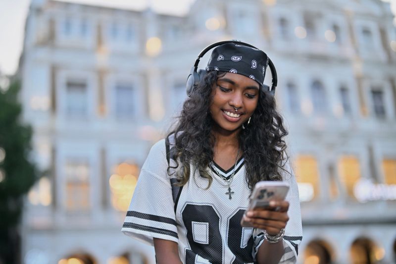 A young girl wearing headphones sits casually on a railing, focused on her smartphone. Her relaxed pose and stylish streetwear capture a candid moment of modern urban youth culture.