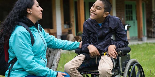 A woman and a boy in a wheelchair share a joyful moment outdoors.