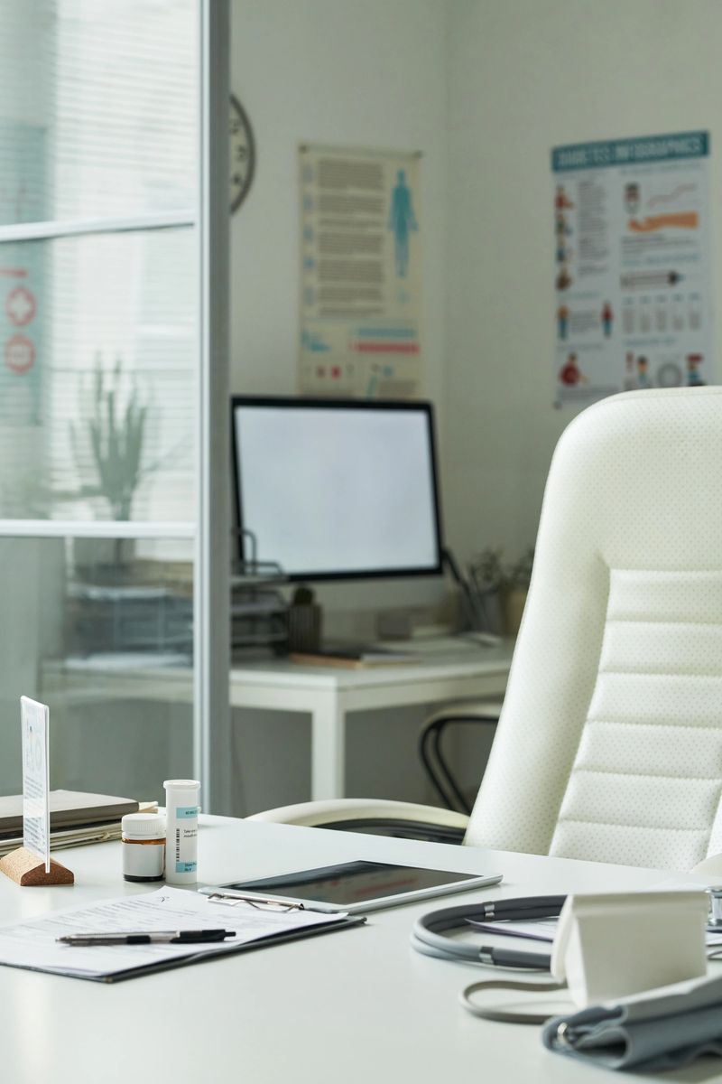 Empty medical office showing desk with blood pressure monitor, prescription bottles, clipboard with documents, computer monitor and posters on wall, representing healthcare setting