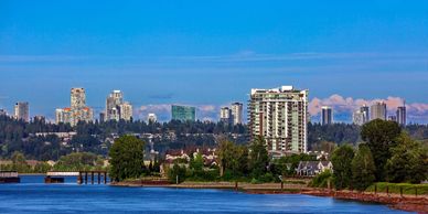 City skyline with modern buildings and a river in the foreground under a clear blue sky.
