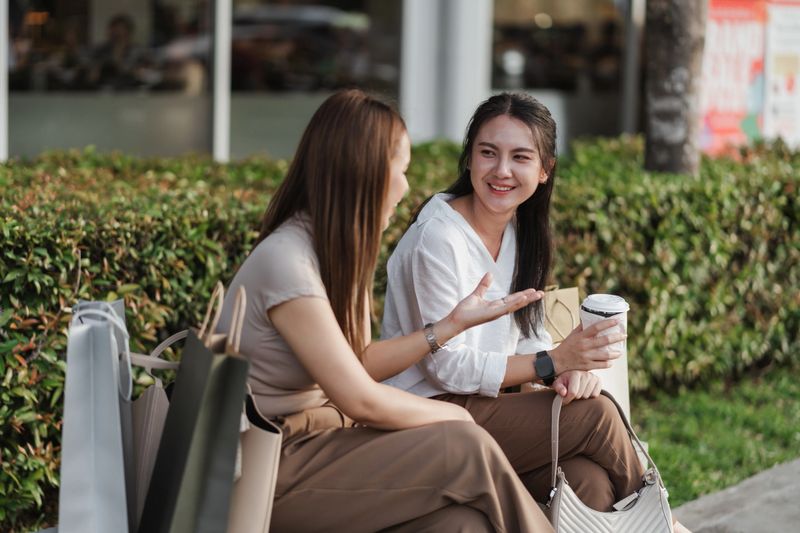 Two Asian women relax on a bench after shopping, smiling and chatting with coffee and bags beside them. A candid lifestyle moment showcasing friendship, leisure, and modern consumer culture.