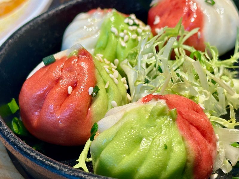 A close-up view of vibrant colorful xiaolongbao, featuring red and green hues, garnished with sesame seeds and served with fresh greens, showcasing creative culinary artistry.