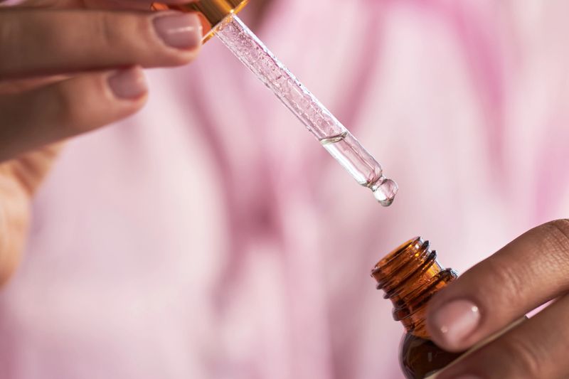 Caucasian young adult woman holding glass dropper above skincare serum bottle preparing for beauty routine, showing close up of hands with natural nails and clear liquid