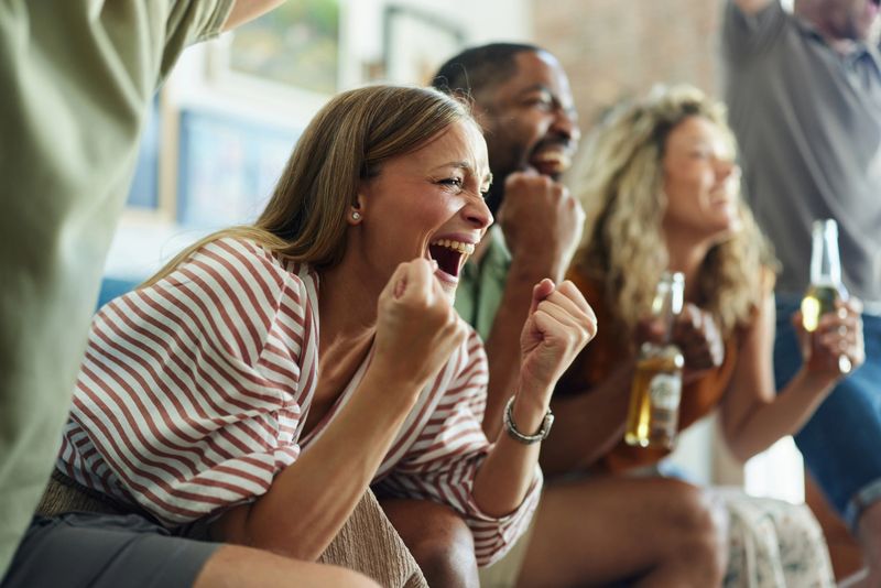 Cheerful woman celebrating the victory of her favorite sports team while watching a match with her friends on TV at home.