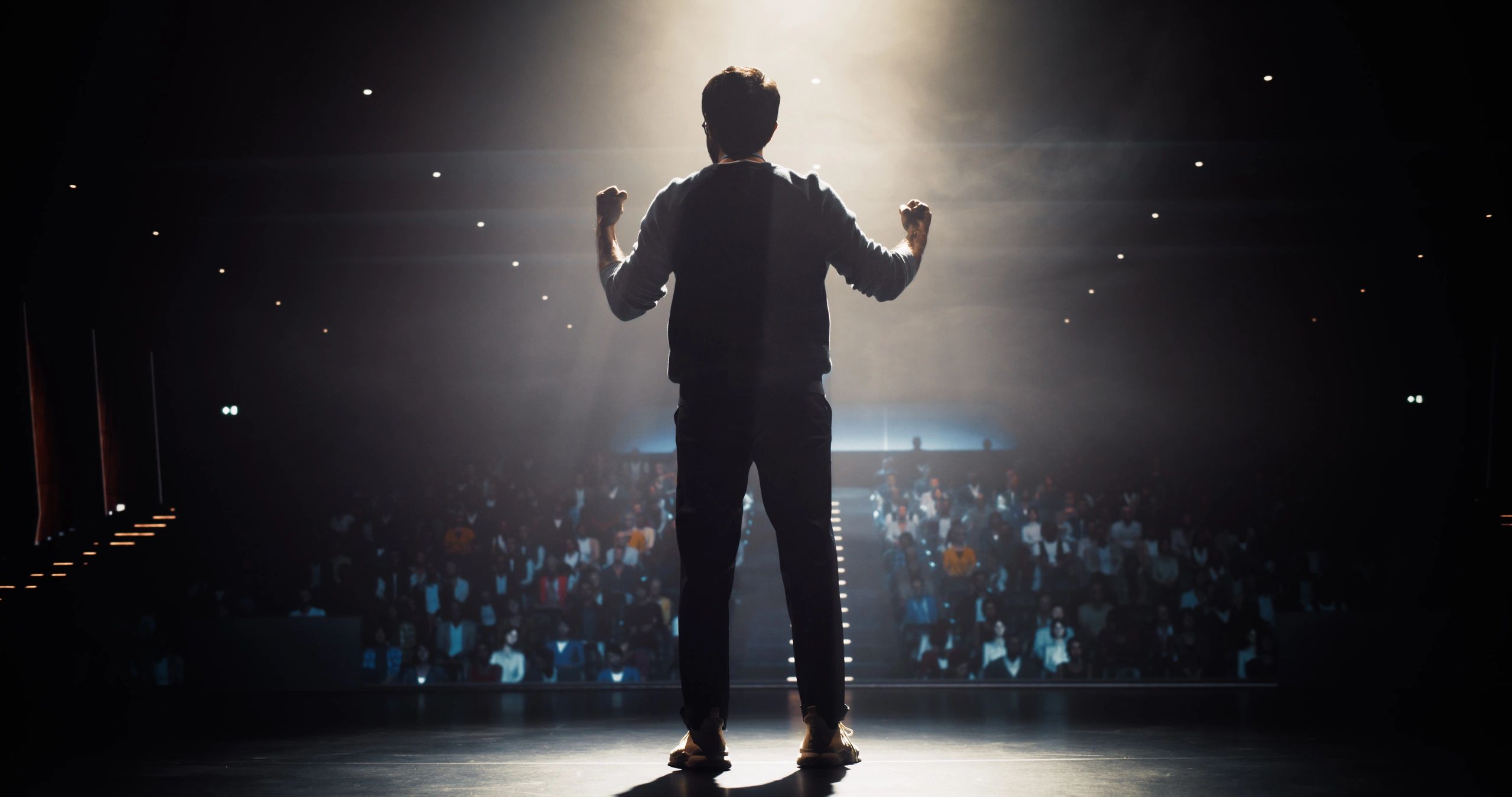 A person stands confidently on stage facing a large audience under bright lights.