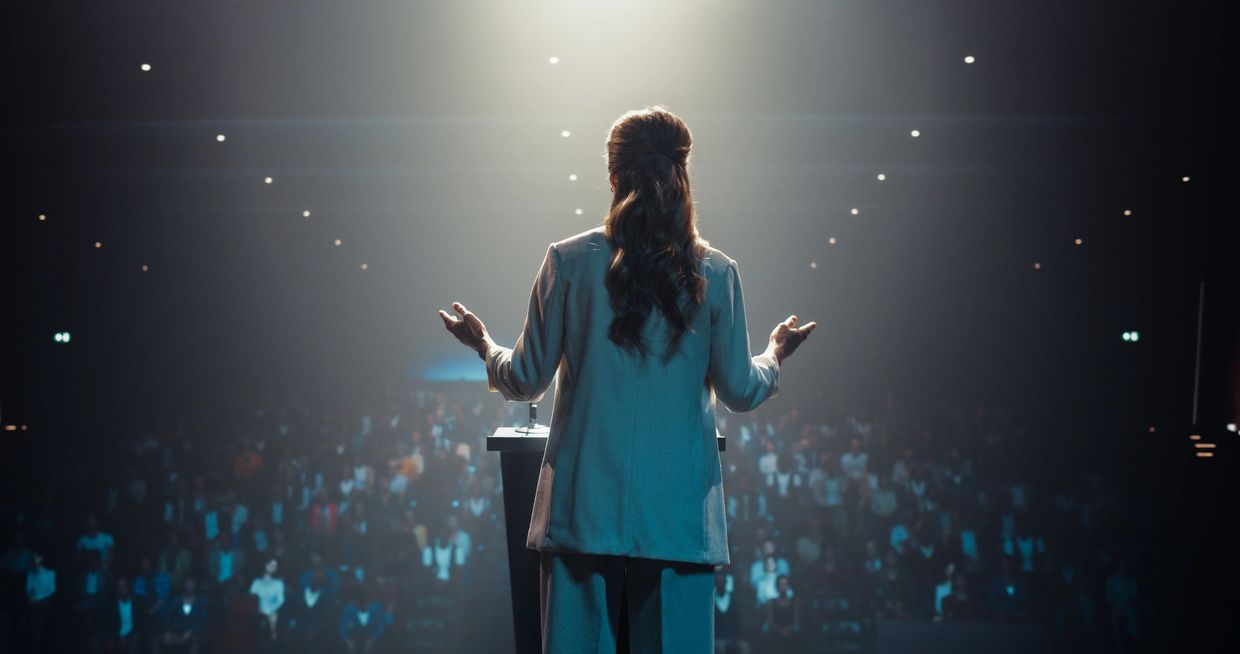 A woman speaking confidently to a large audience in a dimly lit auditorium.