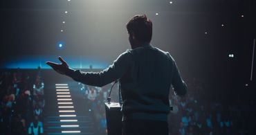 Man speaking to an audience on a stage with dramatic lighting.