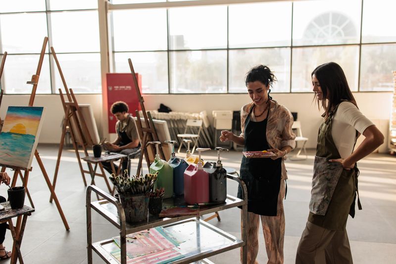 Two women artists in an art class prepare for their next masterpiece. One mixes vibrant colors on a palette while the other artist observes, ready to begin her painting session in the creative environment.