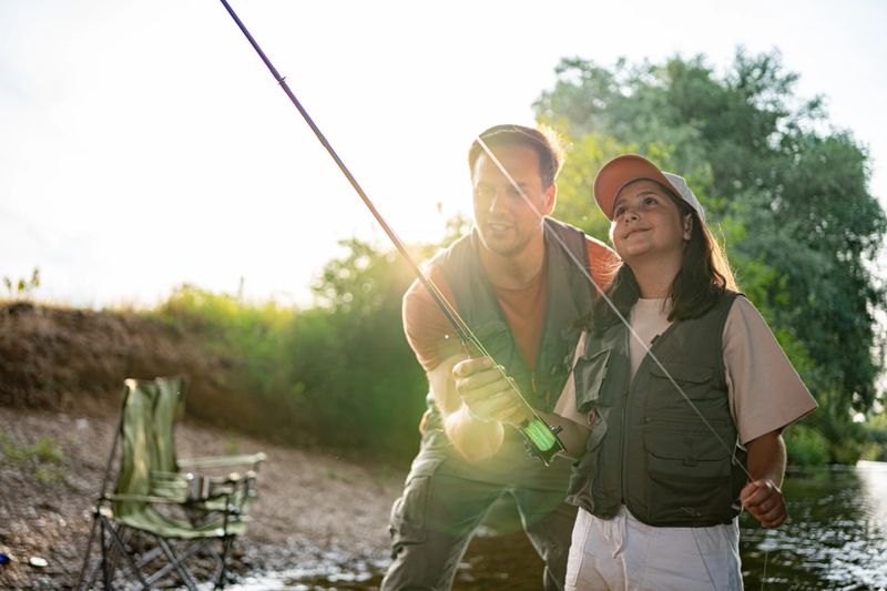 A father teaching his young daughter how to fly fish in a serene river setting. The two are enjoying a sunny day outdoors, bonding while trying to catch trout using specialized fishing techniques.