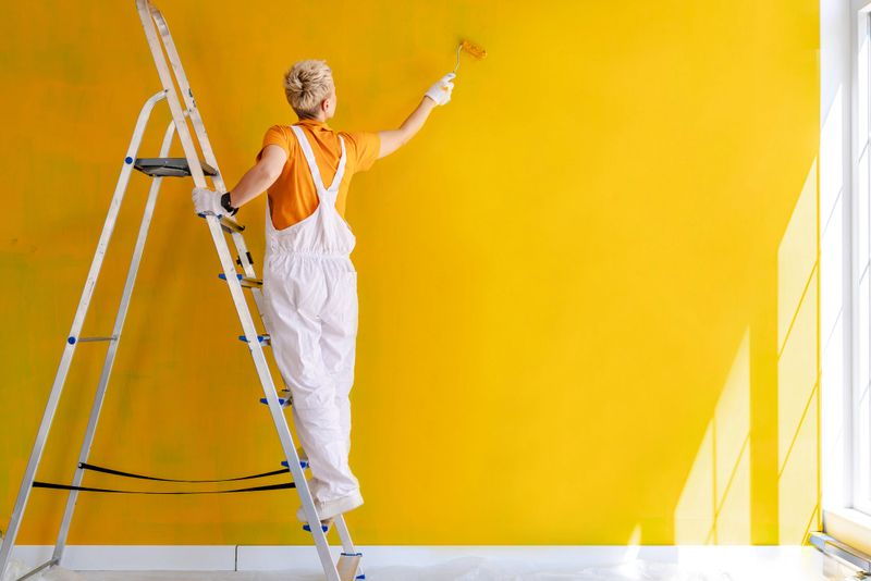 A mid adult woman in white overalls paints a bright yellow wall with a roller while standing on a ladder. She is focused on her craft, enhancing the home interior's aesthetic.