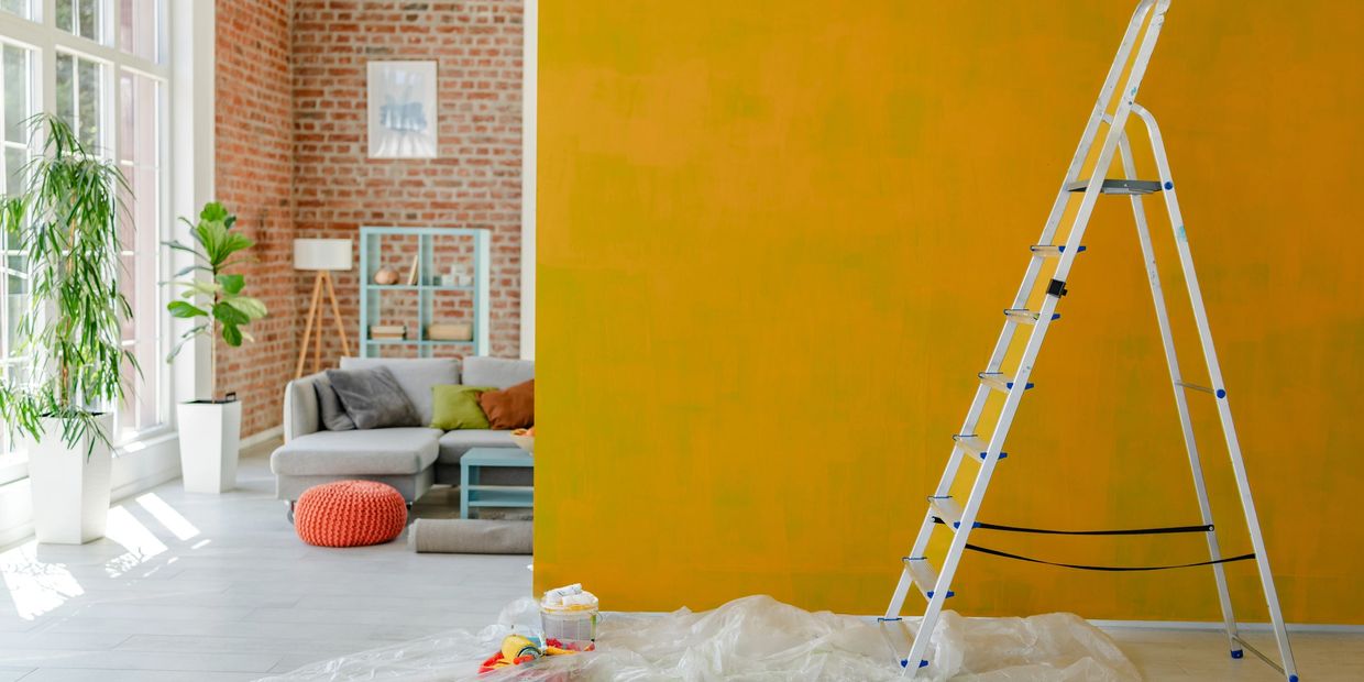 A partially painted yellow wall with a ladder and a cozy living room in the background.