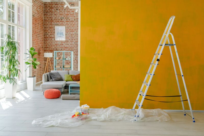Ladder placed beside a vibrant yellow wall mid-paint, with a comfy living space and decorative shelving visible behind.