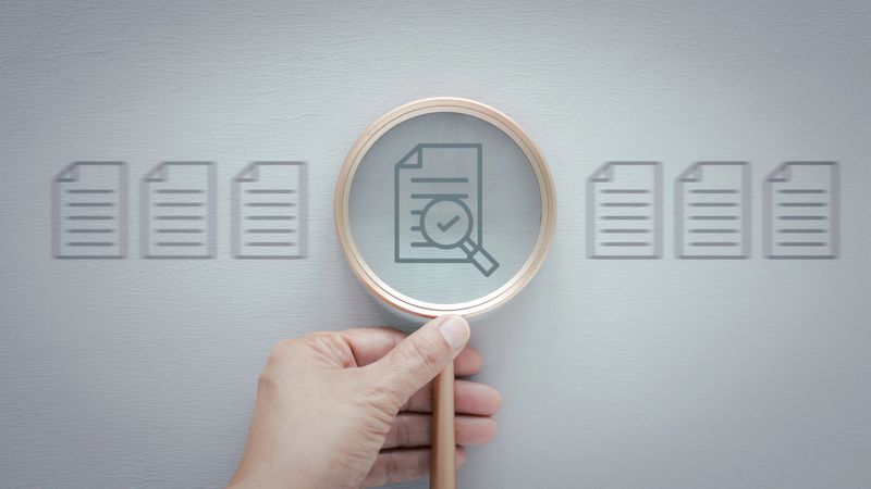 A hand holds a magnifying glass over a document icon that features a checkmark, suggests verification, approval, inspection, or selection over grey wooden background.